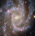 A spiral galaxy seen face-on. Its centre is a bright glowing yellow. The galaxy’s spiral arms contain sparkling blue stars, pink spots of star formation, and dark threads of dust that follow the arms.
