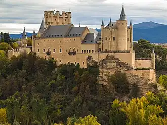 A castle high on a rocky peninsula above a plain. It is dominated by a tall rectangular tower rising above a main building with steep slate roof. The walls are pink, and covered with a sculptural pattern. There is a variety of turrets and details.