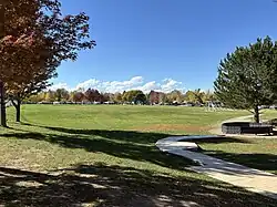 This photograph looks north across the central green at Peak to Peak Charter School in Lafayette, Colorado, with a sinuous concrete path leading from the foreground, shaded edges beneath mature maples and a pine, an open multiuse turf area with scattered worn goalmouths, and cars and trees along the campus perimeter under a bright autumn sky.