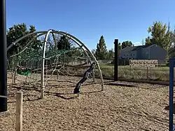 This photograph shows the elementary playground on the Peak to Peak Charter School campus in Lafayette, Colorado, with an arched metal-and-rope climbing frame and molded plastic slide at center, engineered wood-fiber safety surfacing, chain-link fencing at the perimeter, and a colorful anti-hate banner along the east fence under clear afternoon light.
