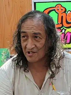 Middle-aged man with long dark wavy hair and light-colored shirt speaking in front of a brightly colored poster