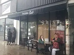 Two wikimedians pose sitting down in front of a pizzeria restaurant, while two others stand underneath a single umbrella to the left/west of the restaurant entrance, on a rainy day.