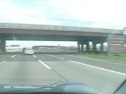 A large bridge hovering a 12-lane wide highway viewed from a car