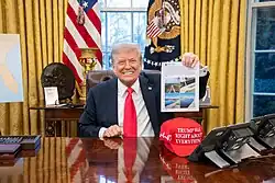 Donald Trump is seated at the Resolute desk and is smiling at the viewer while holding up a printout of a social media post in his left hand. The post has four images of water infrastructure on it.
