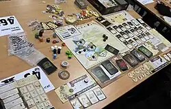 Photograph of the board game set out on a table showing the game pieces, cards and board