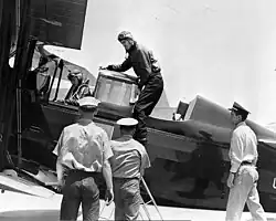 Black and white photograph of three men in khaki uniforms standing in front of a plane's centre, one man in aviator's uniform is deboarding while another person also in uniform is seated in the plane
