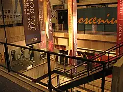 A multi-story interior atrium, made of concrete and with a staircase running downwards in the middle. Several multi-colored banners hang next to the stairs. The back wall has text slightly blocked from view by the banners, with the visible portion reading "osceniu" in cursive.