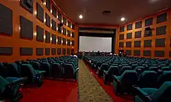 In a lit room, dark square panels line the pinkish wall in rows, while green seats fill the space; a gray screen is visible to the front.