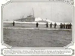 Black and white photo of a ship sinking into the water, with multiple people running towards it.