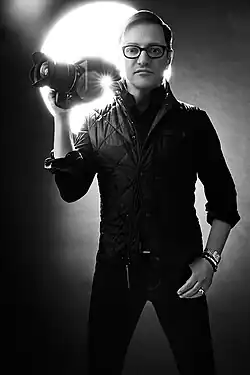 Man wearing glasses and dark quilted vest holds professional camera beside his face, backlit by bright studio light