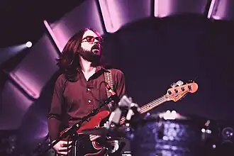 Roy Mitchell-Cárdenas wearing a dark shirt, playing bass guitar on a darkened, purple-tinged stage, with eyes partly closed and mouth slightly open