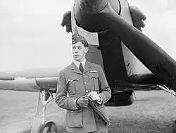 A black and white photograph of the head and torso of a man in uniform, wearing a side cap and gloves, standing in front of a fighter aircraft