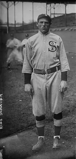 A black-and-white picture of a man wearing a baseball uniform standing on a baseball field with other players and the stadium grandstand in the background