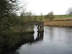 Brackish-looking water with a high wall rising out of it (part of an old mill) with sloping fields beyond