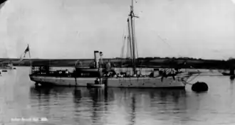A small ship lies moored to a buoy; the ship has a single thin mast and a short smoke stack, and several sailors can be seen aboard.