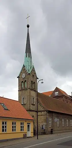 A gothic style church with a tower. The tower is three stories tall and has a copper roof, topped with a sphere and a cross for a spire. The length of the church is parallel to the road, and has a red roof and white windows. The church is built close to a small, residential, yellow house. The sky is overcast.