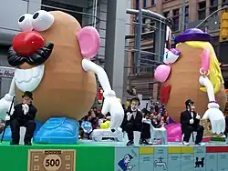 A smiling potato toy with light blue shoes, blue pants, white arms, a brown mustache, a black derby hat, pink ears, an open mouth with lips, a red, round nose and round Pac-Man shaped eyes