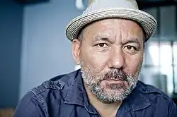 Portrait photograph of a man with a gray beard wearing a blue shirt and tan hat