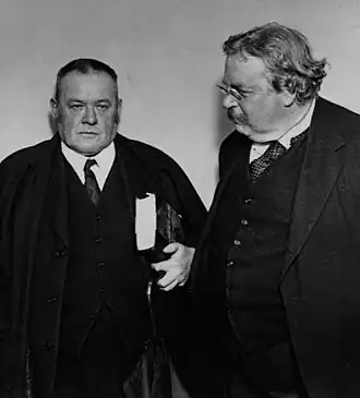 Two middle-aged men in suits and ties; the man on the left has sideburns and cropped hair and is holding a large book and an umbrella in his left hand; the man on the right is obese and has unkempt hair, a moustache, and small glasses