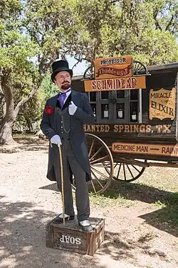 An old-timey man with a long moustache is wearing a suit and tophat and standing on top of a soapbox. Behind him, there is a caravan with adverts describing his snake oil.