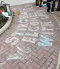 "Stirchley welcomes all with love" is written in bold letters with multi-coloured chalk on a brick pavement, with multi-national flag bunting and a "Hold on to hope" poster in the background