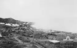 A beach crowded with military stores. Near the foreshore a plume of smoke rises
