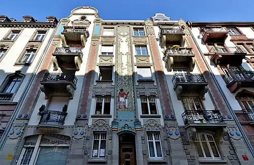 Ornate apartment building facade with balconies and decorative reliefs, with central painted panel of ancient Egyptian figure among stylized lotus motifs