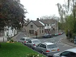 Street scene showing grass area to the left of road junction. Ahead are buildings.
