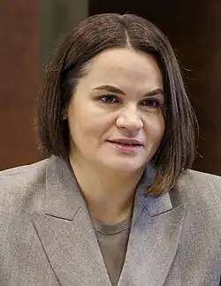 A woman speaking with mid-length brown hair, earrings, and a beige suit