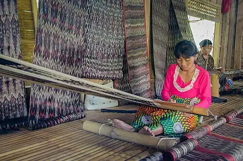 T'boli dream weavers using two-bar bamboo backstrap looms (legogong) to weave t'nalak cloth from abacá fiber. One bar is attached to the ceiling of the traditional T'boli longhouse, while the other is attached to the lower back. The cloth is being patterned by dying the warp, so the loom equipment is simple; a heddle rod, a shedding stick, and a batten. She is also using a footrest. Philippines.[42][43]