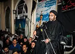 Shia Muslims attending a Majlis in Kashmir, with a cleric delivering a sermon at the microphone during Muharram observances.
