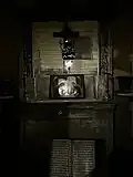 The room is dimly lit, and the focus of the scene is a framed icon resting upon an old stone altar. The icon shows Mary, Joseph, and the infant Jesus a depiction of the Holy Family glowing softly under a hanging lantern.