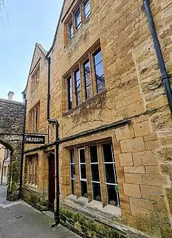 Exterior view of the Abbey Gatehouse in Sherborne, a three-storey historic building faced in warm Ham Hill stone, with mullioned windows and a hanging sign reading ‘Museum.’ An arched stone passageway is attached on the left, and the street runs alongside the building.