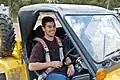 A man in his early thirties is smiling and sitting inside a yellow tomcar and strapped inside by a seatbelt