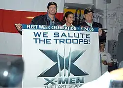 Hugh Jackman, Halle Berry and Kelsey Grammer hold a flag with the X-Men: The Last Stand logo and the inscription "We Salute Our Troops" in a ship's deck.