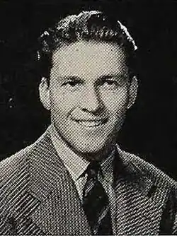 A monochrome bust photo of a white man with dark hair, he is wearing a suit and tie, looking to the camera's right.