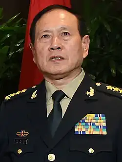 a smiling man with his mouth open and looking rightwards, wearing a light green military uniform with insignia and badges
