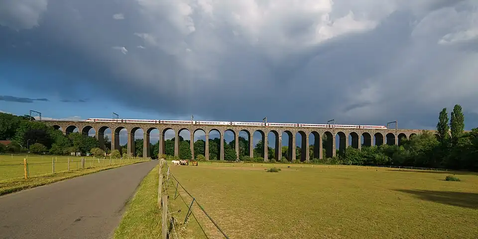 A photograph of an arched brick railway viaduct as taken from the ground quite some distance away. The whole viaduct is in the shot but some is obscured by trees and shrubbery. A red and white express train is passing over the viaduct beneath the metal masts attached to the arches.