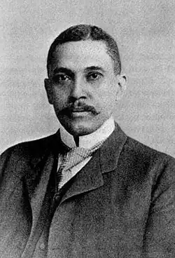 A grayscale head-and shoulders portrait of Wilford H. Smith, a black man with a handlebar mustache and dark hair parted slightly to the left of center. He is wearing a dark suit jacket and vest, a white shirt with a high collar, and a checked tie tucked into his vest.