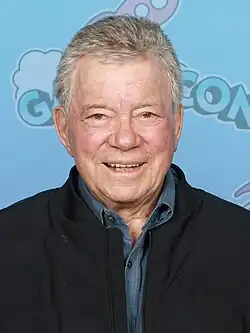 Headshot of William Shatner in GalaxyCon Richmond. He is a later-aged, clean-shaven, light-skinned man with grey hair. He is wearing a button-up shirt and a jacket on top.