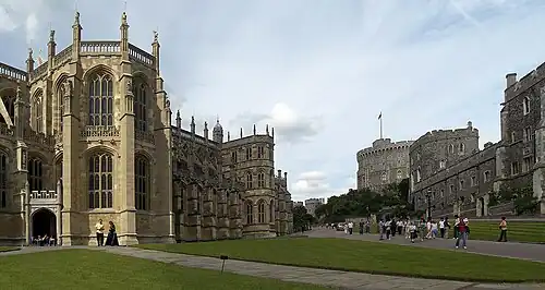A large Gothic chapel on the left, with tall thin windows. On the right is a line of stone buildings, pointing towards a circular tower in the middle of the picture. In the centre are two paths surrounded by grass, with a number of people walking around.