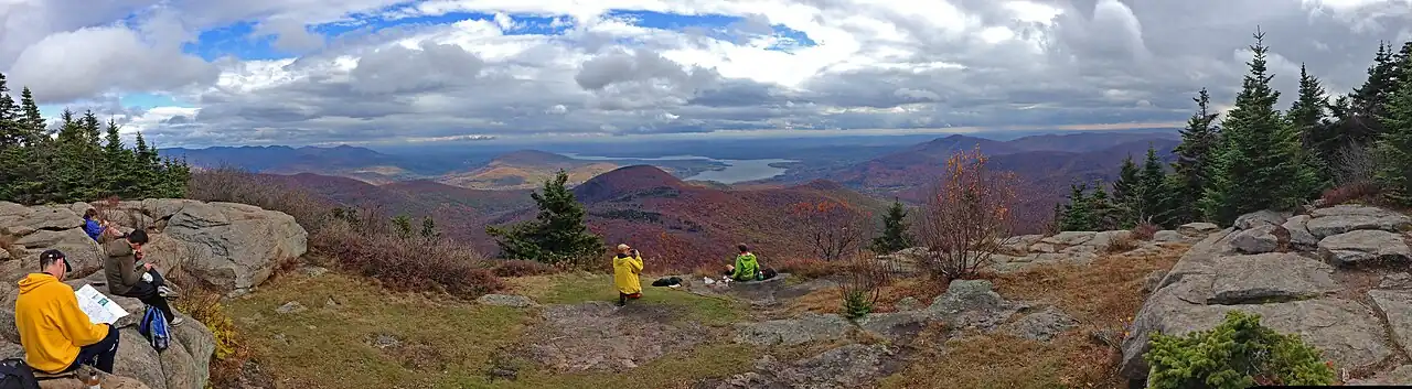 A wide open ledge, with small coniferous trees on either side, on which several people are sitting down and reading, eating or taking photographs. Beyond it lies a mountainous region, with many peaks in fall color. A large body of water is visible just right of center, at the foot of some smaller mountains.
