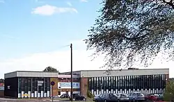 A building approximately 3 stories high. For the most part, the building is made up of horizontal columns of an opaque white panel up to around head height, with 3 windows arranged vertically above this. The columns are approximately 0.5 m (1.6 ft) wide and run from just below the roof to just above ground level and are separated by vertical wooden beams. Above the columns of windows a corrugated white border runs along the top of the building.