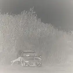 A black-and-white photo of a car parked in front of a hill covered in tall grass.