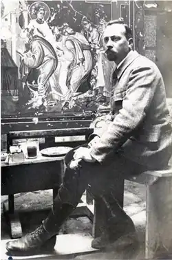 An artist in a jacket, trousers and boots, sitting in front of a Byzantine mural which he is engaged in repainting. He has short, dark hair and a short beard, both neatly groomed.