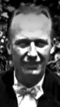 Black-and-white photographic portrait of a smiling man with slicked-back hair and a fuzzy shadow under his nose, plus a sharp one over half of his chin. He dons a bowtie.