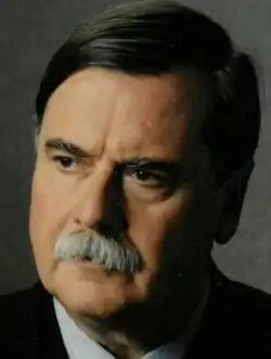 Three-quarter profile, colour publicity shot of an unsmiling man with striking dark eyes and eyebrows, heavy features, a grey walrus moustache, and dark hair with side parting.