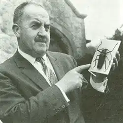 Monochrome, waist-length newspaper photo of a balding man with a moustache. He is wearing a single-breasted suit jacket and patterned tie, and holding and pointing to a pinned specimen or model of a beetle, about four inches (10cm) long in the body.