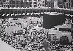 A black-and-white photograph of a large black cube surrounded by people. It sits in the middle of a large courtyard.